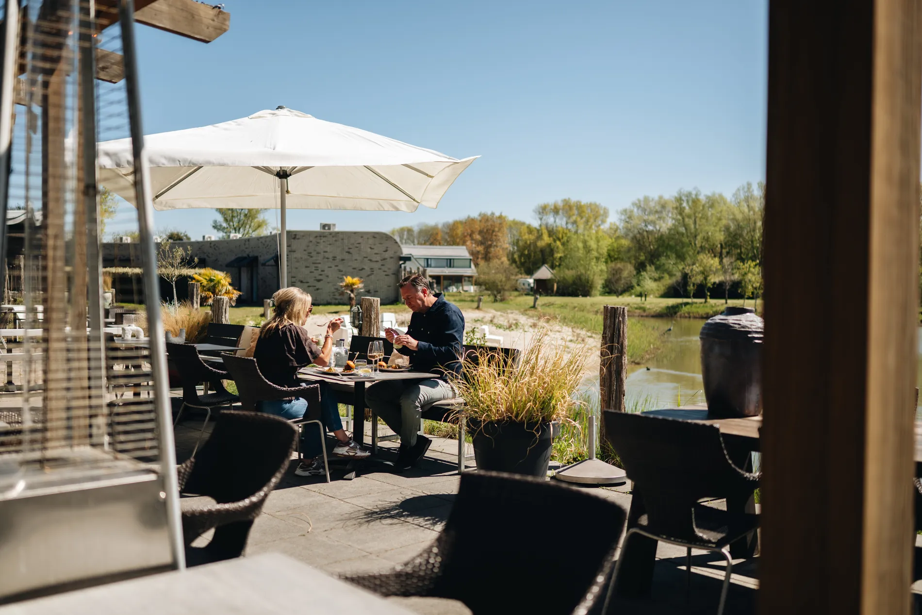 Een man en een vrouw lunchen op het zonnige terras van Restaurant Zeeuws Verlangen onder een grote witte parasol, met uitzicht op een rustige vijver en groene natuur.